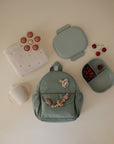 A mushie Flower Press Toy, a light green backpack with wooden beads, a white blanket with small red designs, a green divided lunchbox with cherries, a separate green container, a white sippy cup, and brown toy rings.