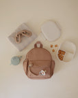 A beige backpack, a striped folded cloth, mushie’s Silicone Pacifier Clip | Hera, a white lunchbox with snacks, and a light blue sippy cup are neatly arranged on a cream surface in minimalist style.