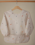 The mushie Long Sleeve Bib, featuring colorful small dinosaur prints, cream color, wide pocket, and machine washable fabric, hangs on a wooden hanger against a neutral background.