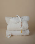 A stack of white towels topped with mushie's Silicone Pacifier Clip | Hera, a food-grade silicone baby bib, and two pacifiers arranged in a minimalist style against a neutral beige background.