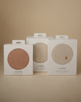 Three boxes of mushie Stretchy Crib Sheets in minimalist packaging sit side by side on a beige background, each with a circular cutout showing the sheet color inside: muted brown, off-white, and beige.