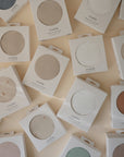 Several boxes of mushie silicone placemats in neutral and pastel shades sit flat on a light surface, each box revealing a round placemat—perfect to pair with the mushie Stretchy MINI Crib Sheet.