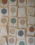 A set of mushie Stretchy MINI Crib Sheets in assorted colors, laid out flat on a beige surface. Each breathable sheet is packaged in a white box with a circular cutout revealing the color inside.