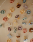 The FRIGG Daisy Natural Rubber Pacifier 2-Pack features pastel daisy-shaped pacifiers and clips in blue, pink, yellow, and brown, each with a natural rubber latex nipple, by FRIGG.