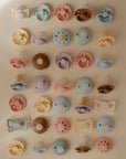 A neatly arranged grid displays FRIGG Daisy Natural Rubber Pacifier 2-Packs in pastel shades, featuring daisy-shaped FRIGG pacifiers with natural rubber latex nipples in various shapes on a light background.
