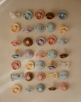A grid of 25 FRIGG Moon Natural Rubber Pacifier 2-Packs, featuring pastel colors and unique designs by FRIGG, is arranged neatly on a beige background—ideal for baby essentials.