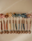 Ten FRIGG Daisy Natural Rubber Pacifier 2-Packs with pastel flower and butterfly designs, featuring beaded silicone and wooden clips, are arranged in a row on a beige surface.