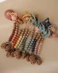 A row of mushie Silicone Pacifier Clip | Eva, crafted from food-grade silicone and wood, is arranged on a beige surface. Each pacifier clip features unique color combos with wooden beads and circular clips at the end.