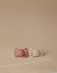 The FRIGG Lucky Symmetrical Silicone Pacifier 2-Pack (6-18 Months) by FRIGG features two medical grade silicone pacifiers, one pink and one cream, with butterfly-shaped shields, displayed side by side on a beige background.