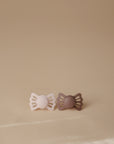The FRIGG Lucky Symmetrical Silicone Pacifier 2-Pack (6-18 Months) features one light pink and one brown pacifier made with medical-grade silicone nipples, supporting healthy oral development. Both are placed side by side on a beige background.