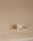 The FRIGG Lucky Symmetrical Silicone Pacifier 2-Pack (6-18 Months) features light brown and cream pacifiers with medical grade silicone nipples for healthy oral development, displayed side by side on a beige surface.