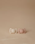 The FRIGG Lucky Symmetrical Silicone Pacifier 2-Pack (6-18 Months) features one off-white and one light pink pacifier, both with medical-grade silicone nipples, displayed side by side on a beige surface against a neutral background.