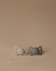 The FRIGG Lucky Symmetrical Silicone Pacifier 2-Pack (6-18 Months) by FRIGG features beige and gray butterfly-shaped pacifiers with medical grade silicone nipples, displayed side by side on a smooth, neutral beige surface for baby comfort.