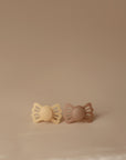 The FRIGG Lucky Symmetrical Silicone Pacifier 2-Pack (6-18 Months) features two butterfly-shaped silicone pacifiers in light beige and light brown, displayed side by side on a smooth, neutral beige background.
