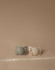 The FRIGG Butterfly Anatomical Silicone Pacifier 2-Pack (0-6 Months) features sage green and cream pacifiers, designed for your baby’s comfort, shown side by side on a soft beige background.