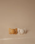 The FRIGG Butterfly Anatomical Silicone Pacifier 2-Pack (0-6 Months) includes mustard yellow and white pacifiers, both with anatomical nipples for comfort, shown side by side on a neutral beige background.