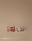 The FRIGG Butterfly Anatomical Silicone Pacifier 2-Pack (6-18 Months) by FRIGG features one pink and one white butterfly-shaped pacifier, shown side by side on a beige background for baby comfort.