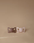 The FRIGG Butterfly Anatomical Silicone Pacifier 2-Pack (6-18 Months) features mauve and light pink pacifiers, placed side by side on a beige surface with a matching background for gentle baby comfort.