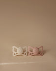 The FRIGG Butterfly Anatomical Silicone Pacifier 2-Pack (6-18 Months) by FRIGG features one white and one pink butterfly-shaped pacifier, shown side by side on a light beige surface with a matching background.
