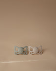 The FRIGG Butterfly Anatomical Silicone Pacifier 2-Pack (6-18 Months) by FRIGG features two pacifiers—one light green, one light beige—with circular centers and butterfly-shaped handles, resting on a neutral beige surface for baby comfort.