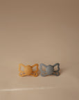 The FRIGG Butterfly Anatomical Silicone Pacifier 2-Pack (6-18 Months) features yellow and gray butterfly-shaped pacifiers side by side on a beige surface, designed by FRIGG for baby comfort.