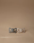 The FRIGG Butterfly Anatomical Silicone Pacifier 2-Pack (6-18 Months) features gray and beige pacifiers with a butterfly-shaped shield, displayed side by side on a light beige surface against a plain beige background for baby comfort.
