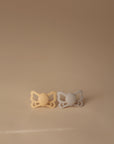 Two FRIGG Butterfly Anatomical Silicone Pacifiers (6-18 Months), one light yellow and one light gray, are placed side by side on a smooth beige surface with a matching beige background.