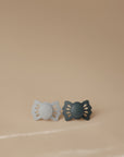 The FRIGG Lucky Symmetrical Silicone Pacifier 2-Pack (0-6 Months) by FRIGG features two medical-grade silicone pacifiers with butterfly-shaped shields in light and dark gray, supporting baby oral development. Displayed on a beige surface and background.