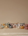 A row of FRIGG Butterfly Anatomical Silicone Pacifier 2-Packs (0-6 Months) in pastel silicone with butterfly-shaped handles is arranged on a soft beige surface, set against a matching background.