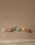 Five pastel FRIGG Lucky Symmetrical Silicone Pacifiers (0-6 Months) with bow-shaped handles, in cream, pink, green, blue, and yellow shades, feature medical-grade silicone nipples for baby comfort and safety. Sold as a 2-Pack.