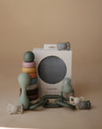 A non-toxic mushie Stacking Rings Toy, silicone teether, pacifiers, and a white mushie box are arranged on a beige surface against a neutral background.