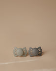 The FRIGG Lucky Symmetrical Silicone Pacifier 2-Pack (0-6 Months) features butterfly-shaped shields in green and gray tones. Each pacifier has a medical-grade silicone nipple, designed for comfort and safety, shown on a beige background.