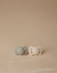 Two FRIGG Lucky Symmetrical Silicone Pacifiers (0-6 Months), one light gray and one cream, with butterfly-shaped shields and medical-grade silicone nipples, rest side by side on a beige surface against a neutral background.