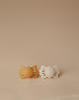 The FRIGG Lucky Symmetrical Silicone Pacifier 2-Pack (0-6 Months) includes one mustard yellow and one white pacifier with bow-shaped handles. Made of medical grade silicone for comfort and safety, both are shown side by side on a beige background.