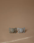 The FRIGG Butterfly Anatomical Silicone Pacifier 2-Pack (0-6 Months) by FRIGG, featuring one green and one light gray pacifier, is displayed side by side on a beige surface, highlighting their comfort-focused design for your baby.