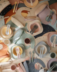 A flat lay of mushie Square Dinnerware Plates, made from BPA-free silicone, arranged in overlapping rows. The plates feature soft pastel shades like blue, pink, yellow, green, and orange.