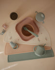 A flat lay of pastel baby feeding items—bowls, spoons, Mushie Trainer Sippy Cup with easy-grip handles, and silicone placemats—all neatly arranged. The non-toxic Mushie cup comes in soft pink, blue, and beige shades.