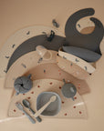 A flat lay of toddler feeding items, featuring the mushie Snack Cup, along with bibs, plates, bowls, utensils, and placemats in soft beige and blue tones with adorable animal and star designs.