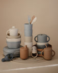 A neatly arranged collection of mushie Snack Cups in spill-proof pastel silicone, plus baby dishes, utensils, and containers for toddlers in soft beige, blue, and taupe on a light surface against a neutral background.