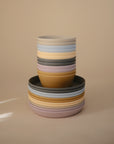 Mushie Round Dinnerware Plates in blush, mustard, sage, blue, and cream are stacked against a beige background. Dishwasher safe and stylish, these plates add practicality and charm to everyday meals.