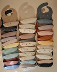 A vertical display of mushie Silicone Baby Bibs with adorable animal, rainbow, and flower patterns; each non-toxic bib features adjustable sizing for a perfect fit, neatly arranged against a neutral background.