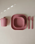 The mushie Dinnerware Cutlery Set includes a pink silicone sippy cup with handles, two stackable plates, spoon, and fork—all non-toxic and dishwasher-safe—arranged on a light beige background.