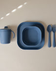 The mushie Square Dinnerware Plates set, made from blue BPA-free silicone, includes a sippy cup, bowl, fork, and spoon—all displayed neatly on a beige surface.