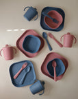 Four mushie Dinnerware Cups sets for children, each featuring a plate, bowl, fork, spoon, and non-toxic cups in solid blue and pink. All items are displayed on a neutral background.