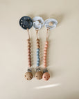 Three mushie Hera Silicone Pacifier Clips are displayed side by side on a light beige surface, each with a minimalist gray, white, and light blue design featuring earth-toned wooden and food-grade silicone beads.