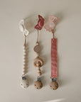 Three mushie Silicone Pacifier Clip | Hera designs are shown: two with minimalist wooden beads, and one with a pink silicone ribbon. Each has butterfly or flower shapes and secure food-grade silicone holders.