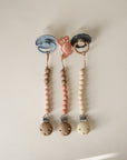 Three Hera silicone pacifier clips by mushie are arranged in a row on a light background. The blue, pink, and black pacifiers feature neutral-toned beads and minimalist design made from food-grade silicone for safe, stylish comfort.