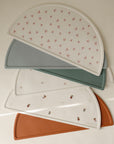 Four mushie Silicone Placemats in muted tones, made from food-grade silicone, are stacked diagonally. Each placemat displays a minimalist pattern with flowers, geometric shapes, or tiny figures.