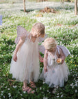 Two young children in white fairy dresses and wings stand among white flowers, one pointing to the ground as they radiate comfort and calm—just like FRIGG Butterfly Anatomical Silicone Pacifier 2-Pack (6-18 Months) from FRIGG. Another child in wings appears behind them.