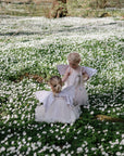 Two children dressed as fairies sit among white flowers, sharing treats from colorful mushie Silicone Baby Food Containers in a sunlit forest clearing.